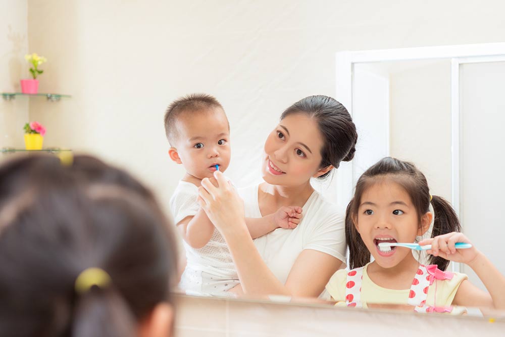 mom holding little baby son using toothbrush cleaning his teeth and teaching cute girl daughter